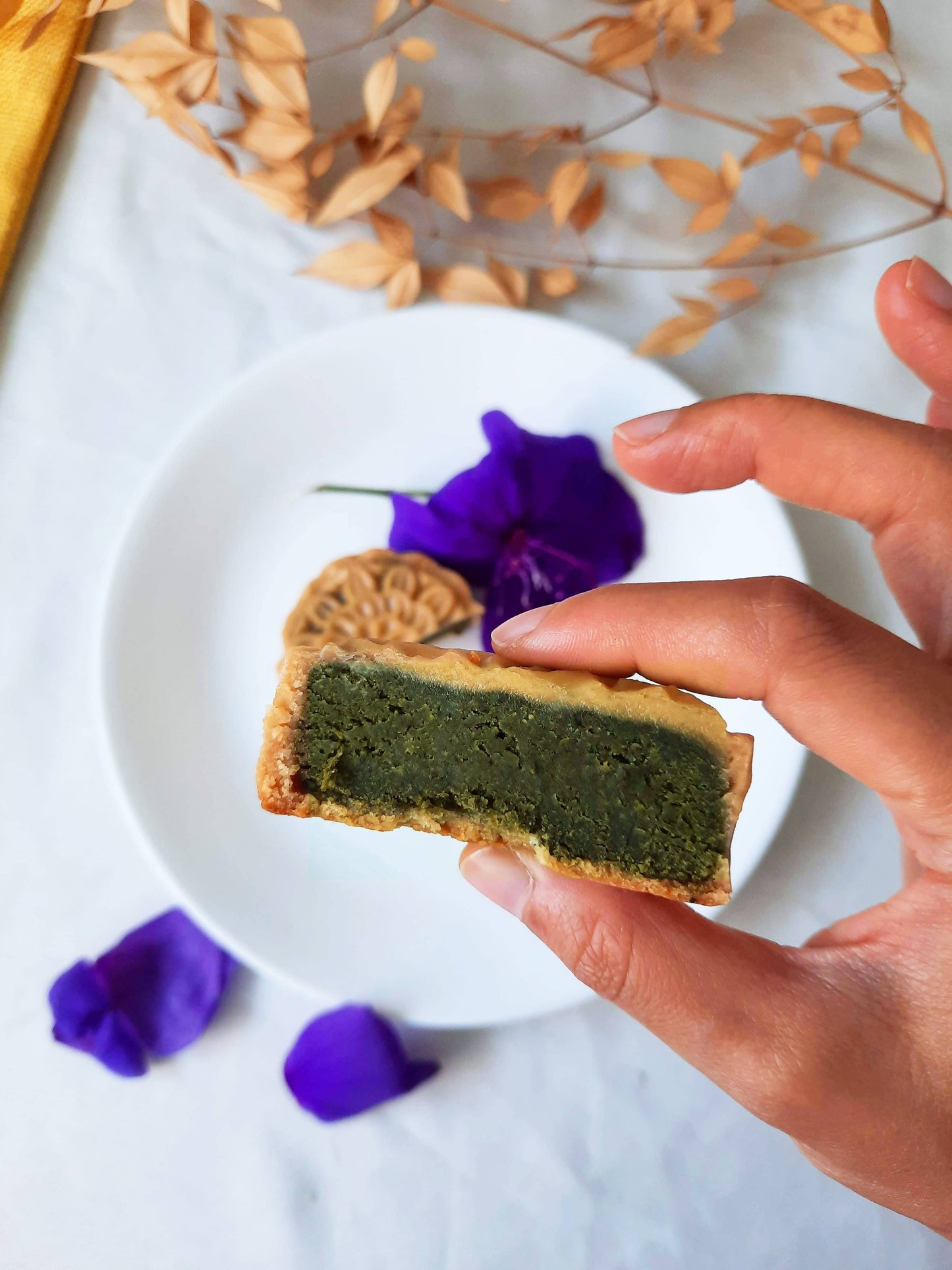 Hand holding up half a jasmine tea mooncake towards the camera. Jasmine tea filling showing to the camera. Background is the second half of the mooncake with leaves and purple flower petals.