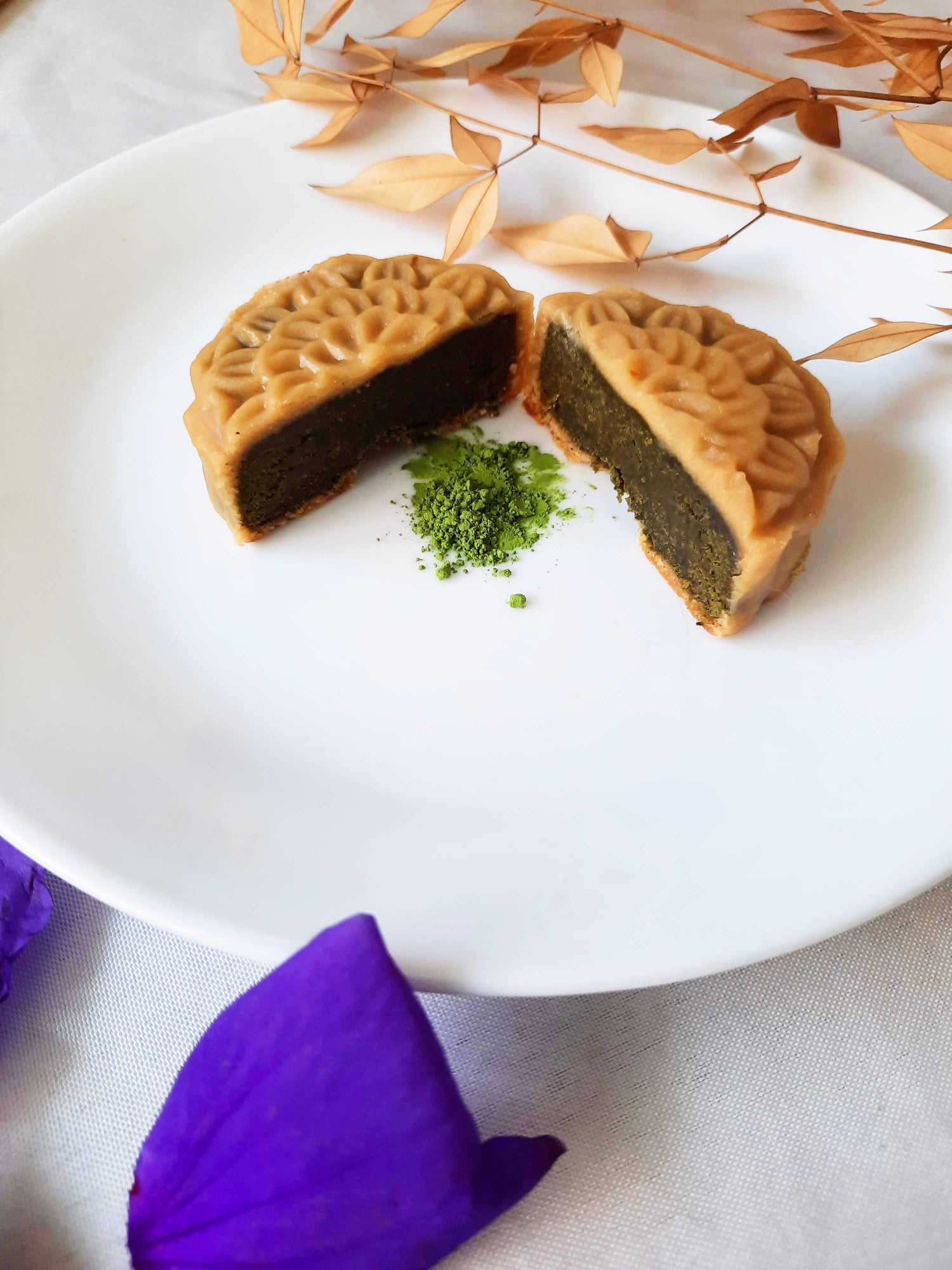 Matcha mooncake cut in half with matcha powder in between the two halves. Matcha filling showing the the camera. Purple petals towards the forefront of the camera with leaves on a thin branch in the back.