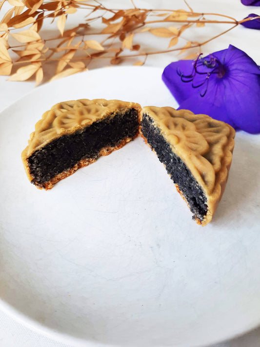 Black sesame mooncake cut in half with filling shown to camera. Background is white with purple flowers and brown leaves on a thin branch.