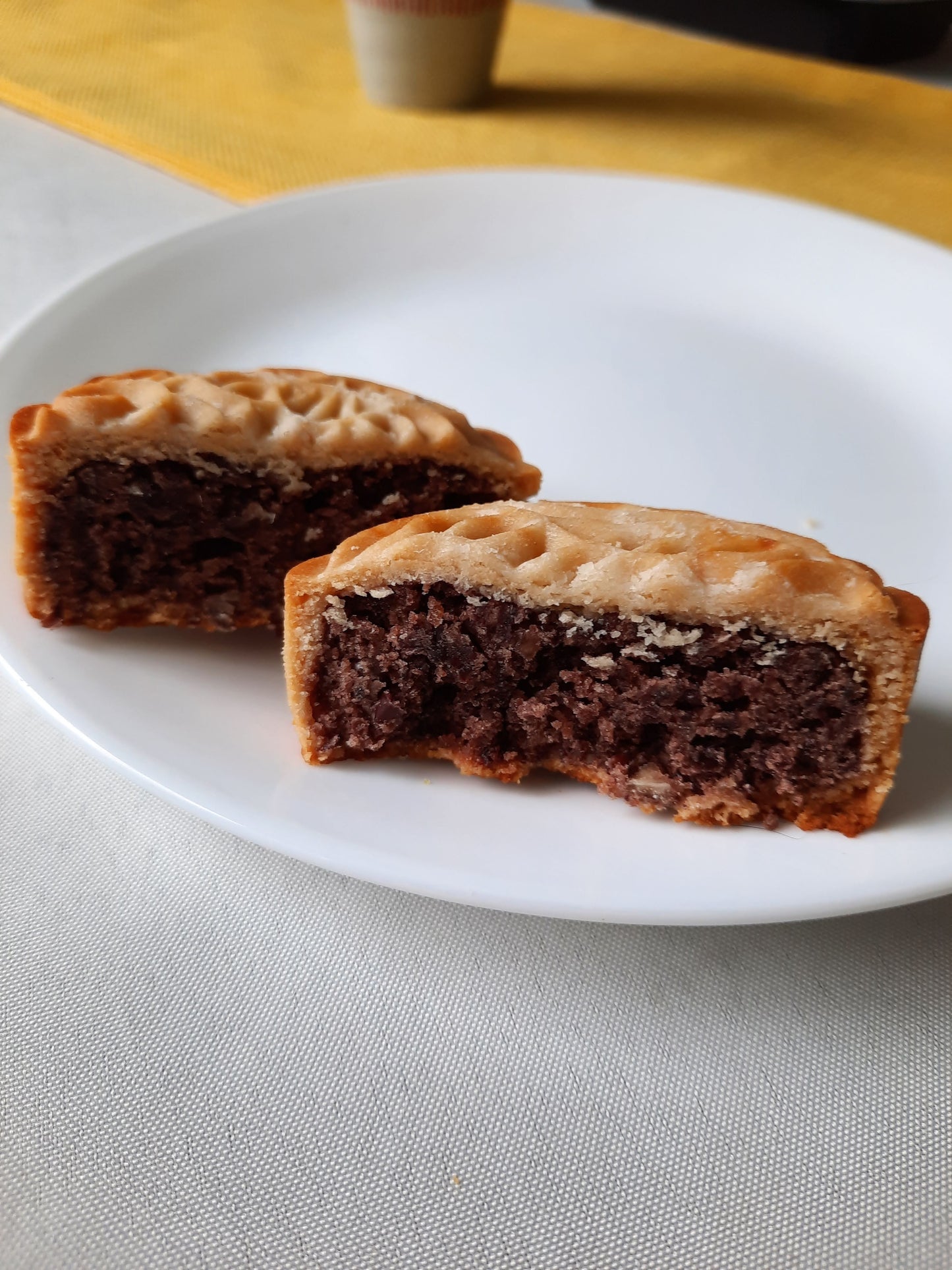 Two halves of a red bean mooncake on a white plate with red bean fillings facing the camera. A cup of green tea on a yellow cloth in the background.