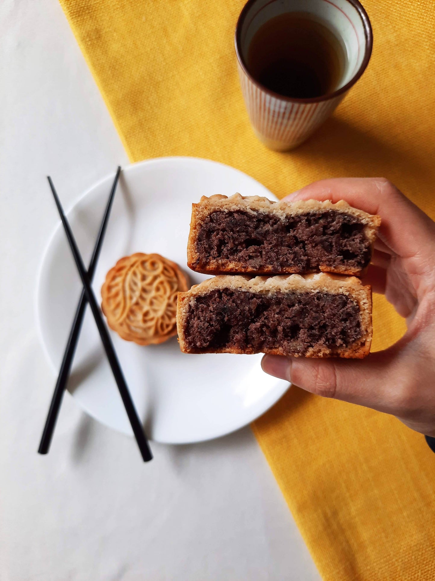 Two halves of a red bean mooncake stacked and held up to the camera with red bean filling showing straight to the camera. In the background is an overhead view of a full, round red bean mooncake on a plate with chopsticks to the left and a cup of tea to the upper right corner. 