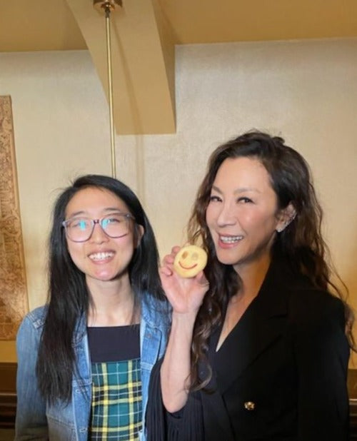 A picture with maker Annie Wang alongside Michelle Yeoh. Yeoh is dressed in a black suit with flared sleeves and is holding up the smiley faced almond cookie made by Annie's T Cakes. Picture was taken at the premier of Everything Everywhere All At Once.
