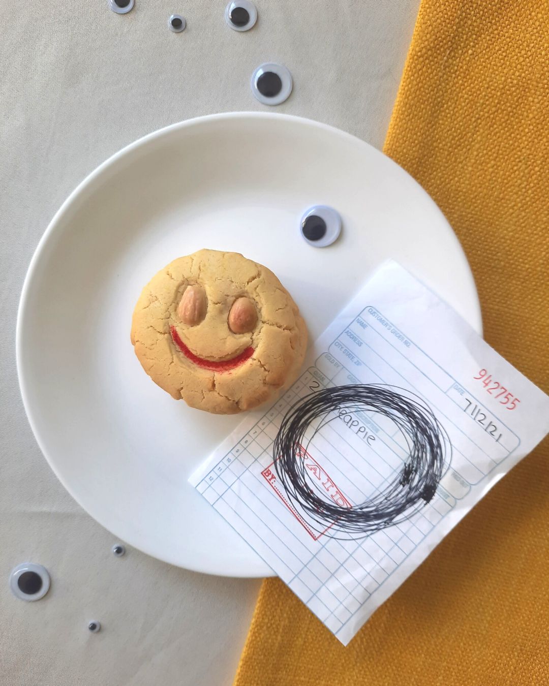 A smiley faced almond cookie with red lips on a plate. Next to the cookie is a receipt with a wild circle drawn in the middle. The foreground has different sizes of googly eyes.