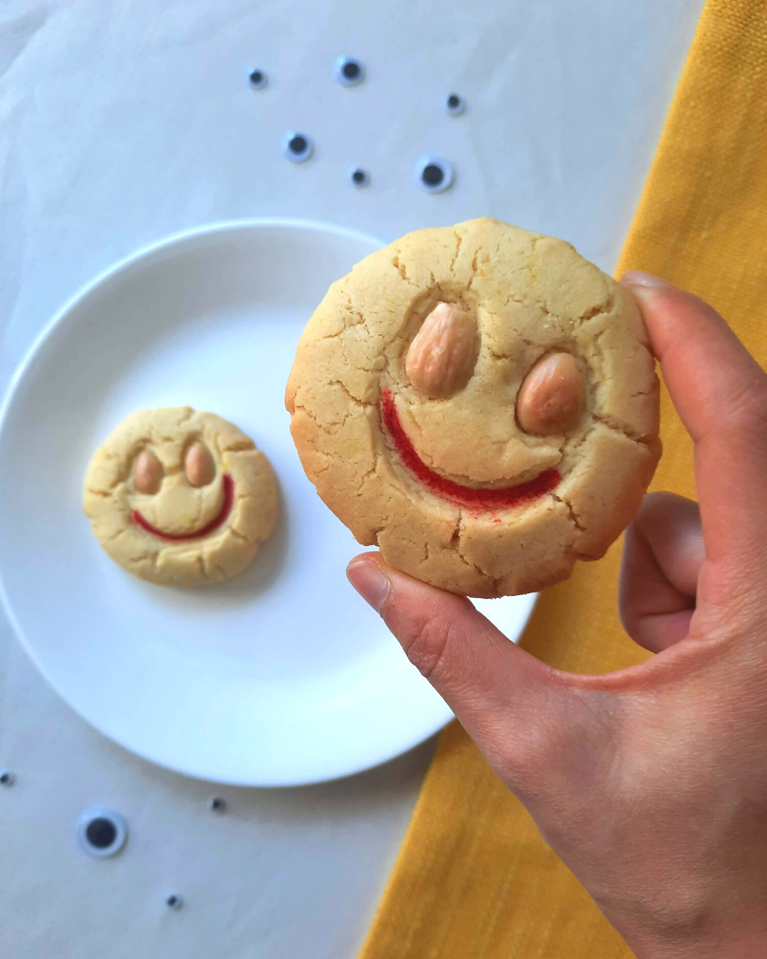 A hand holding up one smiley almond cookie with red lips in front of the camera. Another cookie on the table below the foreground almond cookie.